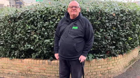 Nigel Briers stands in front of a low wall and a bush. He is dressed in a black hoodie and holds a walking stick. 