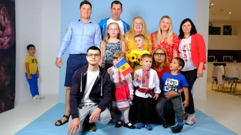 Chiara MacCall A Romanian group, posing for a photo, in a studio, one child is holding a flag, while another is away from the camera standing in a corner