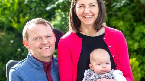 My Name'5 Doddie Foundation Scott is on the left and is smiling in the picture, his wife is sitting beside him and is wearing a pink cardigan and black top. She has brown bobbed hair and is smiling. Their baby is on Robyn's lap.