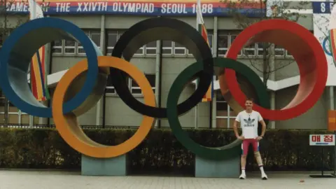 Mark Williams Mark as a young man stands in front of a large statue in Seoul, South Korea, showing the five Olympic rings. He has close-cropped blond hair and is wearing a white T-shirt with a large Adidas logo on the front and tight cycling shorts in Team GB colours of red, white and blue.