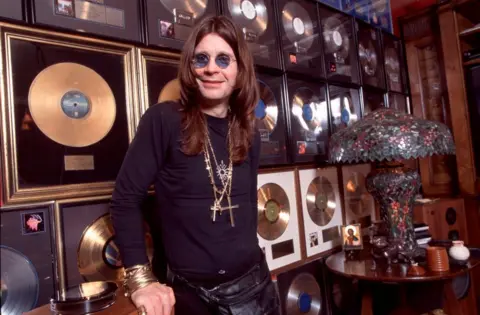 Getty Images Ozzy Osbourne wearing round dark glasses and gold necklaces stands in front of a collection of gold and platinum discs