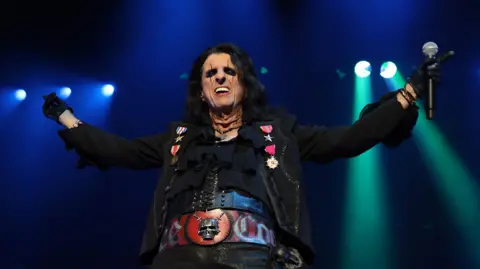 Rock star Alice Cooper on stage with his arms thrown in the air, a microphone held in one hand. He has black hair, heavy black eye makeup, black clothes, and a thick red belt with a silver skull on it.