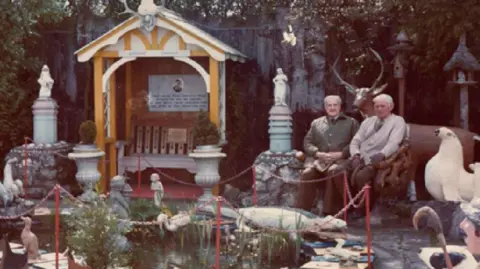 James and Christina Beveridge Two men with grey hair sit among a garden with dozens of cement animals and creatures