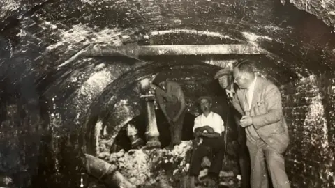 Cambs Times/NewsQuest An old black and white photo showing the inside of an arched brick tunnel, about five foot high in the middle section. Three workmen can be seen, all wearing flat caps and there is one man on the right in a suit, shirt and tie and no hat.