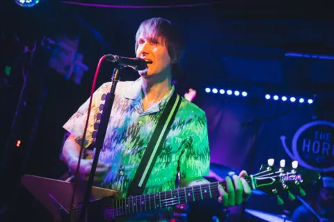 Luke Hinton A man is standing on stage under a spotlight. He is holding a guitar and the words "The Horn" are on the wall behind him.