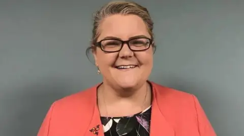 Clare barber, she has long blonde hair that is tied up and is smiling. She is wearing dark-rimmed glasses and a salmon jacket over a black top.