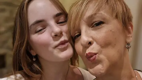 Marianne and her daughter who both look like they're blowing a kiss with their eyes shut. Her daughter has brown bobbed hair and Marianne has short fair hair. They are both wearing dangling earrings.  