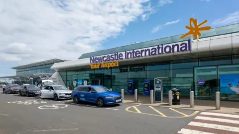 The glass-fronted entrance to Newcastle International Airport. Several taxis are parked outside.