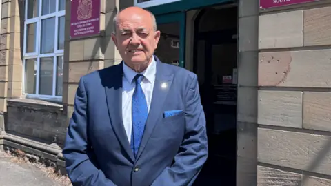 A man in a blue suit and a blue tie with a white shirt. He is smiling but his eyes are slightly closed as the sun are in them.