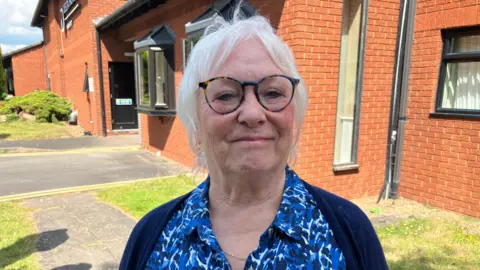 Sam Read/BBC A woman in a blue, white and black shirt with a navy blue cardigan stands in front of a red brick house smiling at the camera.