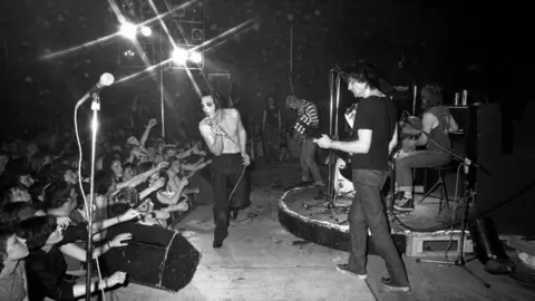 Getty Images Four members of The Damned are on stage. Singer Dave Vanian, who is bare-chested and has his face painted, is stooping at the front of the stage. Many of the audience have their arms outstretched towards him.
