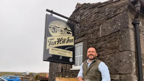 Andrew Hields A man in a white shirt and dark green padded waistcoat is smiling at the camera. He's standing in front of a dark stone building with a window and a hanging pub sign that reads Tan Hill Inn. To the left-hand side is an outside pub area and parked cars.