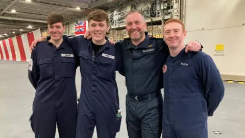 Marc Gaier/BBC Three young men in navy boilersuits and an older man stand on a big ship with their arms around each other. 