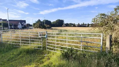 The sun is glinting off a metal gate that encloses a large field. Houses can be seen around the edge and there are bushes and trees.