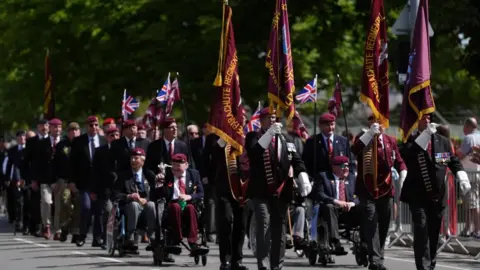 PA Media Four men in suits parade the colours (flags) of the parachute regiment in the road in front of veterans
