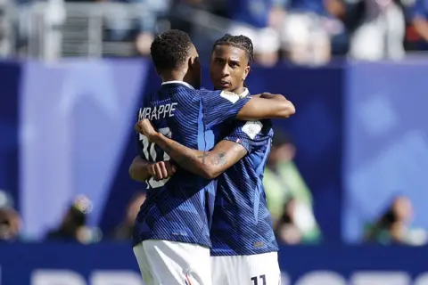 Michael Olise of France (right) and team-mate Kylian Mbappe hug in celebration of France's 3-1 win over Senegal in the 2026 World Cup. Both players are wearing white shorts and navy shirts and Olise is looking at the camera