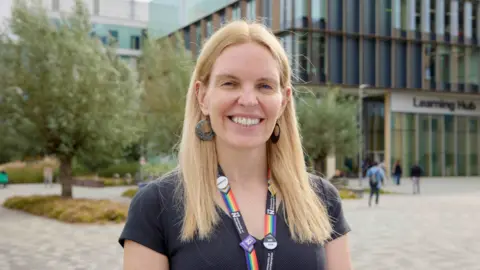 UON Prof Anne-Marie Kilday, standing outside a building, wearing a lanyard, with long blonde hair, smiling and looking at the camera. She is wearing a blue T-shirt and large circle earrings. There are trees in the distance.