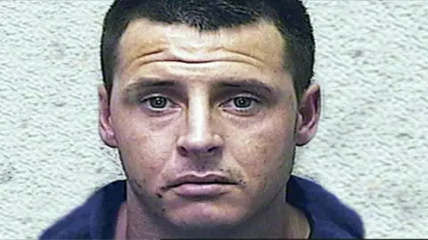 South Wales Police Police headshot of Jason Shaddick. He is a white man in his 20s with close cropped dark brown hair and green eyes. He is wearing a blue top. He is unsmiling and looking straight at the camera.