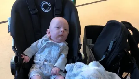 Supplied Robin, as a baby, is pictured in a black car seat. He is wearing a long sleeve top and has dungarees on.