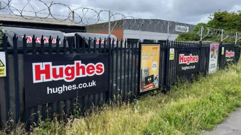 Qays Najm/BBC Black railings-style fencing adorned with Hughes' posters and advertising is in front of an area where trucks  with the Hughes logo are parked. Barbed wire has been fitted to the top of the railings. A grass verge is in front of the fence.