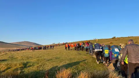 Masses of young people are walking a trail that winds up the top of a tor on Dartmoor. The terrain has large tufts of grass and pieces of granite scattered across it.