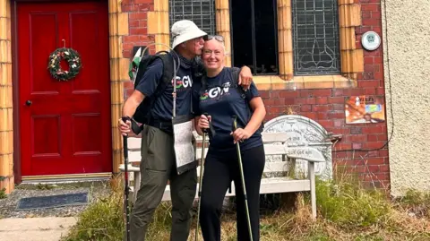 Andy Hockey John kisses Hayley on the forehead, with his hand around her shoulder. They hold walking sticks, and stand in front of a red brick house with a red door.