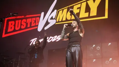 Getty Images Charlie Simpson and Matt Willis from Busted stand on a stage, both with their right arms in the air. They stand in front of a screen that says "Busted VS McFly Tour 2025".