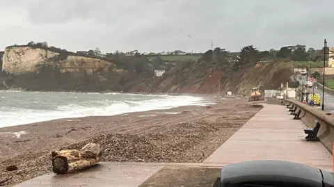 East Devon District Council Seaton beach with shingle on the beach and a clear esplanade. The waves are crashing onto the beach. There is a land in the background and a cliff face.
