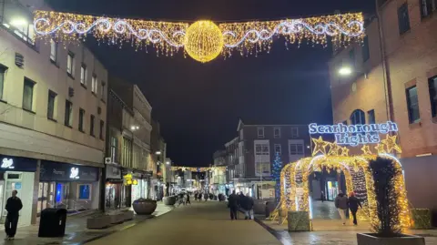Festive street at night with glowing overhead light displays, a bright archway reading “Scarborough Lights,” and people walking past shopfronts reflecting the decorations.