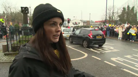 Lucy Williams, wearing dark beanie hat and jacket, speaking as crowds line street behind her