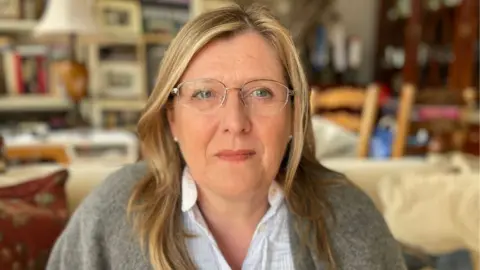 A woman with blond hair and glasses look at the camera while sitting on a sofa in a living room. She is visible from her chest and wears a light -colored shirt under a gray cardigan.