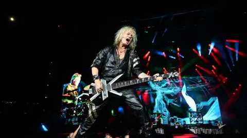 Paras Griffin/Getty Images Joe Elliott is playing guitar on stage while singing. He is dressed in all black leather and has medium length white hair. The stage is dark with some bright blue and red lights shining across it.