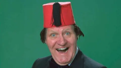 Tommy Cooper grins at the camera wearing a fez in front of a plain green background 