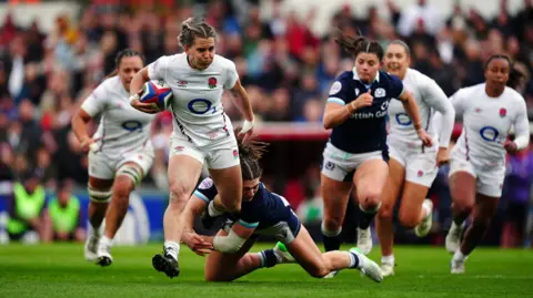 PA England's Claudia MacDonald gets away from Scotland's Helen Nelson to score a try during the Guinness Women's Six Nations match at Mattioli Woods Welford Road Stadium. MacDonald is carrying the ball in her right arm and is puffing her cheeks out as she shrugs off a tackle by Nelson, who has nearly wrapped her arms around her legs. Players from both team are chasing MacDonald.