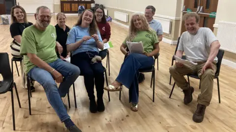 Eight members of the cast of the Whodunnit Theatre Company sit on chairs reading scripts and smiling. They are in a large room with a wooden floor