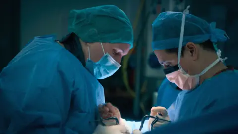 BBC/ExpectationTV Two surgeons operating on a patient in a hospital theatre. They are wearing blue surgical masks and gowns.
