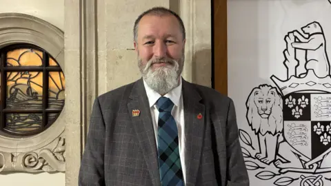 Perry is wearing a grey checked suit and blue tartan tie. He has two badges on his lapels. He has a grey beard and moustache and short hair.