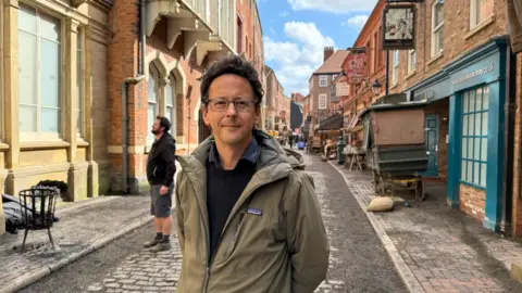 A slim man with dark-brown hair smiles as he stands in a cobbled street which has been dressed up to look like Victorian London with old signs, stalls and street furniture. He is wearing rectangular glasses and a green coat over a black jumper. Another man stands behind him looking towards a grand-looking building. 
