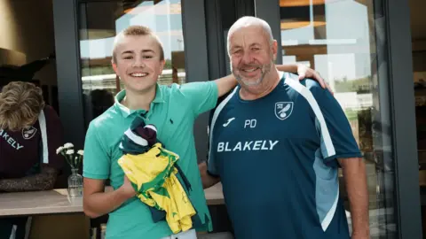 John Fairhall/BBC Ranulph is presented with the Norwich City kit by the club's kitman, Pete Dye. They have broad smiles as they pose for the camera arm in arm with one of the players behind them. Ranulph is holding his new yellow Norwich shirt and maroon training top and green shorts close to his chest in his right arm.