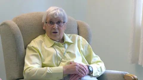 A woman with short grey hair, wearing a yellow blouse sits in a brown armchair