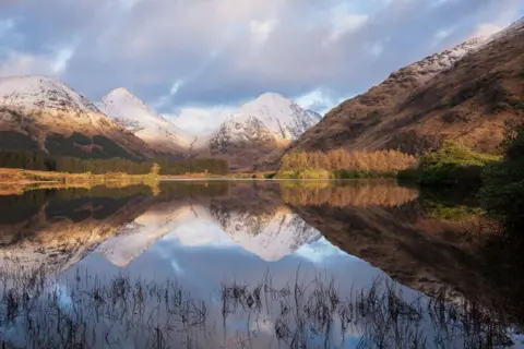 Gwyneth Bell Snow-capped hills, and green/brown trees, reflected in water.