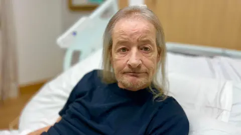 Martin Giles/BBC Gary Jones looking into camera from his bed at the Arthur Rank Hospice. He has long grey hair and grey stubble. He is wearing a navy t-shirt.