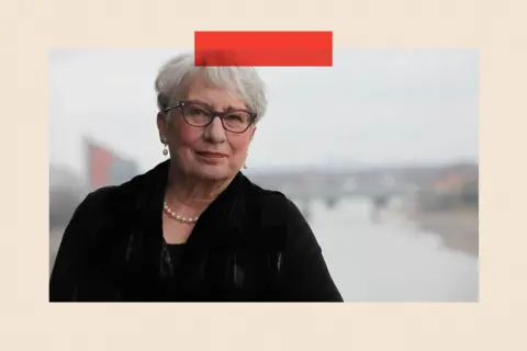 Professor Dame Uta Frith standing in front of a river. She has short grey hair, glasses, pearls and is wearing a dark coat