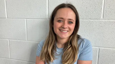 Saffron Cresswell stands against a white brick wall wearing a blue shirt