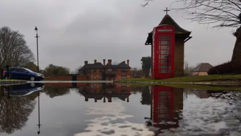 Georgeta A puddle in the road reflects a red phone box, a brick building and a blue car. The photo is a mirror image of the scene with the water stretching across the road. The cloudy sky is grey. 