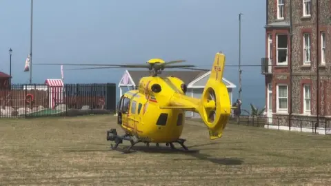 A yellow air ambulance helicopter is sitting on grass beside apartments. Some mini golf holes are located behind a black fence in the background, and behind that, is the sea and the sky.