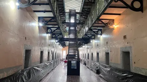 A view of an empty prison landing inside Armagh Gaol. A large, black metal staircase dominates the centre of a prison corridor, with several cell entrances lining either wall. Swathes of black plastic sheeting have been used to block off access to the cells. Large Georgian windows at the end of the corridor are letting in bright sunlight. A number of red plastic chairs and a table covered in white cloth are partially visible behind the staircase. 