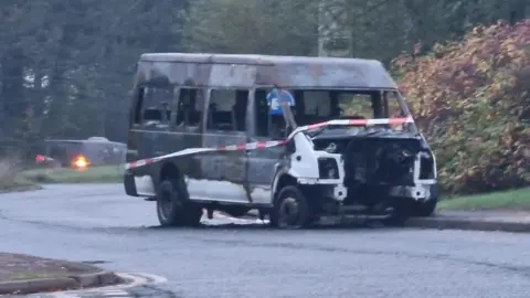 Caroline Haslett Primary School A burnt out bus, with no engine, with tape round it, on a road. A road sign is behind it and buses to the right. The bus was originally white in colour. 