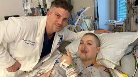 Madi Foster A male doctor wearing a white hospital coat and blue shirt leaning over a woman lying on a hospital bed with tubes and wires attached. She is holding a thumbs up to the camera.