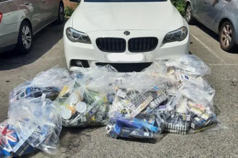 Poole Police Bags of tobacco boxes are piled up in transparent bags in front of a white BMW.